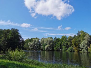 Gölün kıyısı, insanların kayıkla gezdiği yer, ağaçlarla çevrili, güneşli bir yaz gününde bulutlu mavi gökyüzünün arka planına karşı..
