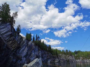 Kayalık sahilin altından bir manzara ve güneşli bir yaz gününde Ruskeala Dağ Parkı 'ndaki Mermer Kanyon' un zümrüt suyuna düşen büyük bir kaya..