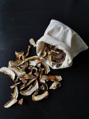 Kurutulmuş porcini mantar ve boletus kapaklarının siyah ahşap bir masaya döküldüğü açık ve sıkıştırılmış bir bez torba manzarası..