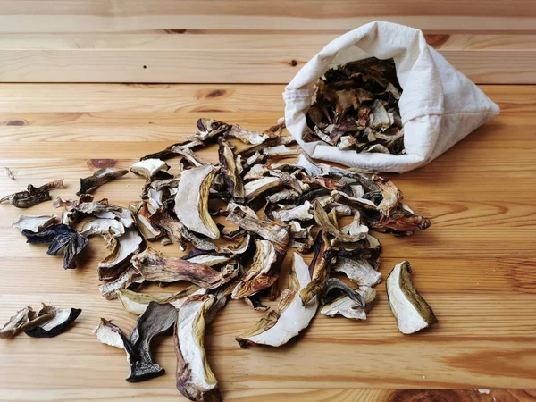 Kurutulmuş porcini mantarları ve beyaz keten bir çantaya saçılmış mantarlar ahşap bir masanın üzerinde uzanır..