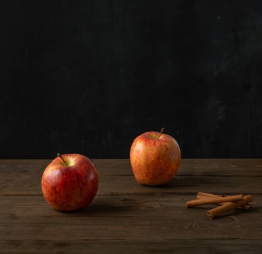 İki kırmızı olgun elma ve tarçın çubukları koyu ahşap bir arka planda.