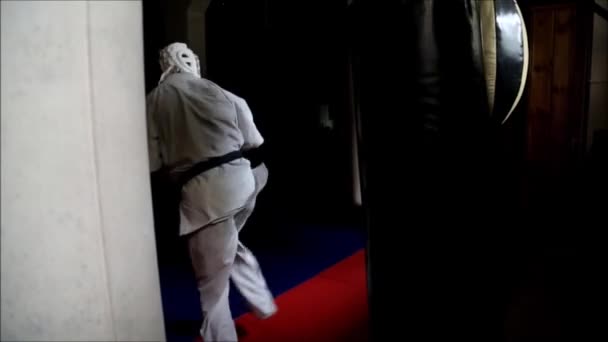 L'athlète en kimono blanc et casque de protection s'entraîne dans le hall sur un sac de boxe. — Video