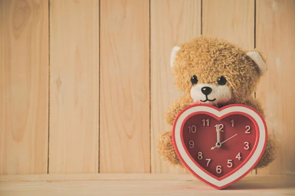 Teddy bear holding red heart clock on wooden table — Stock Photo, Image