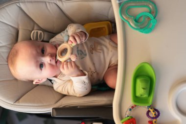 Yükseksandalyedeki Bebek Teether, Minik Ellerle tutulan ahşap yüzük, Silikon Tabak ve Renkli Oyuncaklar Tepside, Yumuşak Tulum, Parlak Mutfak Işığı, Sıcak Ev Atmosferi, Sakin Meraklı