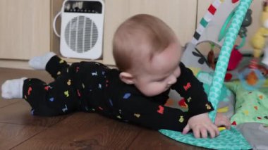 Mat 'te emekleyen, kemer oyuncağına uzanan ve silahlarla iten, konsantre ifade, sert ahşap döşeme ve sıcak ev atmosferi gelişimi gösteren beyaz bebek zamanı.