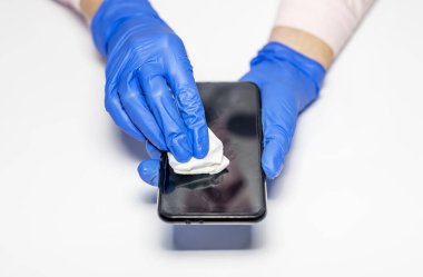 Mavi koruyucu eldivenler giymiş, akıllı telefonu ve cep telefonunu dezenfekte edip temizleyen ve beyaz masaya silen bir kadın eli. Üst görünüm.