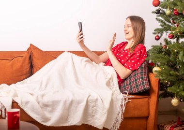 Genç, güzel bir kadın el sallıyor ve cep telefonuyla video konuşmalarıyla sohbet ediyor, süslü Xmas ağacının yanındaki pijamayla kanepede uzanıyor. Evde yalnız yeni yıl çevrimiçi.