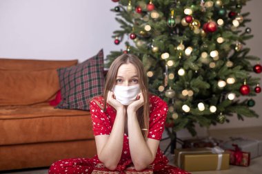 Yılbaşı ağacının yanında oturan genç kadın yüz koruyucu maske takıyor. Kilitlenme ve koronavirüs sırasında evde yalnızdım. Kapat..