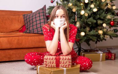 Noel ağacının yanında elinde hediye kutularıyla oturan genç bir kadın yüz koruyucu maske takıyor. Kilitlenme ve koronavirüs sırasında Noel zamanı evde yalnızdım..