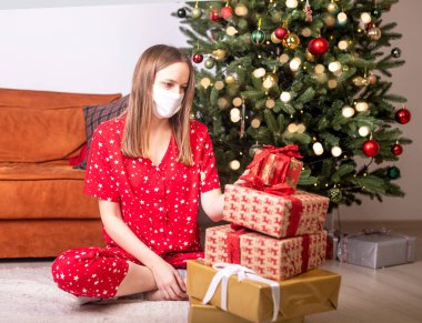 Noel ağacının yanında elinde hediye kutularıyla oturan genç bir kadın yüz koruyucu maske takıyor. Kilit altında ve koronavirüs altında Noel zamanı evde tek başına üzgün..