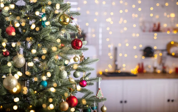 Festive fir tree branches with shiny red and golden baubles, balls, xmas ornaments and bright warm lights, garland. kitchen interior on blurred background.