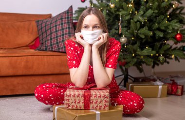 Noel ağacının yanında elinde hediye kutularıyla oturan genç bir kadın yüz koruyucu maske takıyor. Kilitlenme ve koronavirüs sırasında Noel zamanı evde yalnızdım..