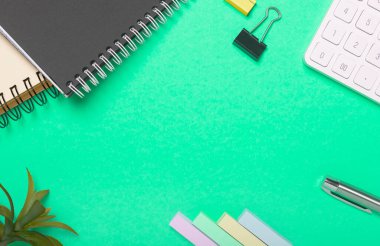 Yeşil ofis masası ya da masa arkası. Arkaplanda bitki, hesap makinesi, kalem ve spiral defter var..