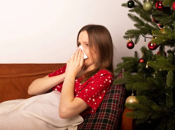 圣诞节时患有流感或头孢病毒的年轻妇女 躺在沙发上 在装饰过的冷杉树旁打喷嚏 — 图库照片