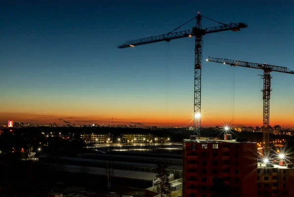 Two construction building cranes over cityscape at evening or night with lights of city.