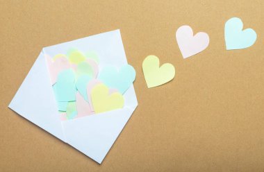 Beyaz zarftan farklı renkte kağıt kalpler kahverengi arka plana saçılmış..