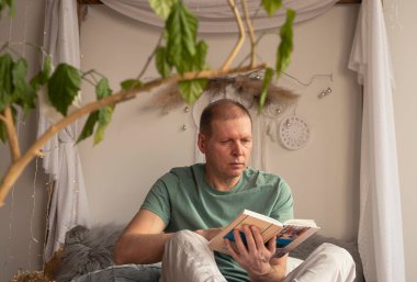 Orta yaşlı yetişkin bir adam rahat bir evde kitap okuyor, gündüz vakti..