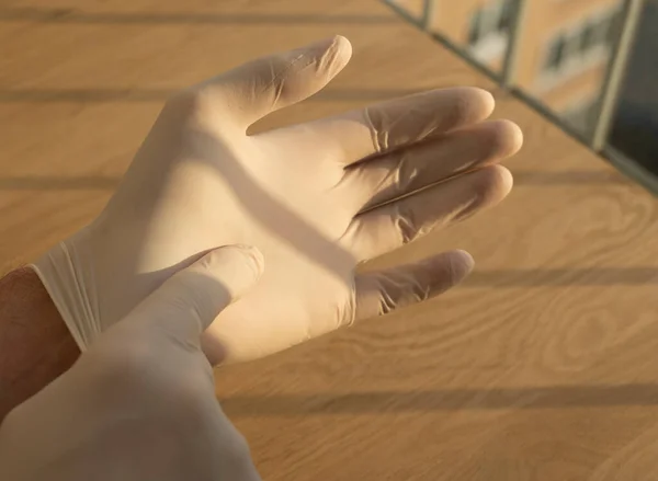 Αρσενικά χέρια λαμβάνοντας καουτσούκ λευκά ιατρικά γάντια πάνω από το ξύλινο γραφείο με το φως της ημέρας από το παράθυρο — Φωτογραφία Αρχείου