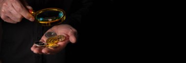 Shiny coins of cryptocurrency in male hand with magnifying glass, over black background, closeup. Banner with copy space clipart