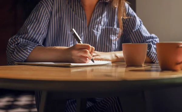 Vrouwelijke hand met pen het nemen van notities, het schrijven van plannen in het dagboek op houten tafel met koffiebekers in koffiehuis. Vrouw aan houten bureau met planner en theebekers close-up — Stockfoto