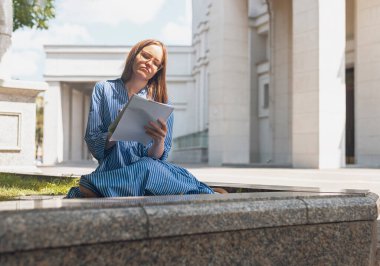 Not defteriyle çimlerin üzerinde oturan, smth yazan, üniversite binasının yakınında notlar alan bir kız öğrenci. Modern kadın kitapla öğreniyor.