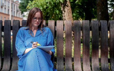 Gözlüklü kadın öğrenci şehir parkında bankta müfredatla ya da fosforlu kalemle oturuyor. Kadın dışarıda çalışıyor.