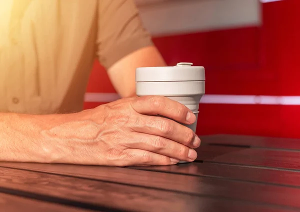Mężczyzna ręce zbliżenie gospodarstwa wielokrotnego użytku krzemu na wynos eko filiżanka kawy w ręce nad drewnianym ulicy stół na dworze — Zdjęcie stockowe