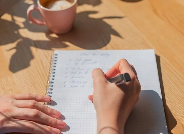 Planos femininos de escrita manual em notebook ou planejador sobre mesa de madeira com luz solar e xícara de café — Fotografia de Stock