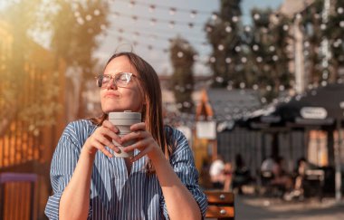 Genç bir kadın sıcak yaz havasının tadını çıkarıyor sokak yemeklerinde kahve fincanı ile dışarıda oturuyor ve güneşe bakıyor ve gün ışığından faydalanıyor, şehir hayatının tadını çıkarıyor.