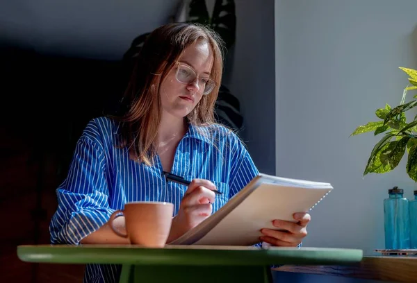 Jovem mulher em óculos escrevendo com caneta no caderno, tomando notas no café com xícara de café na mesa — Fotografia de Stock