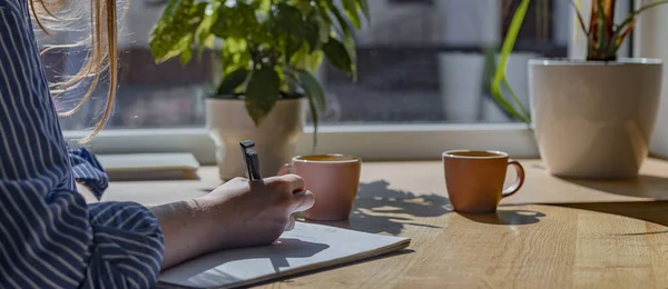 Женщины вручную крупным планом пишут ручкой в блокноте или планировщике, делая заметки в кафе с чашкой кофе и растениями на деревянном столе — стоковое фото