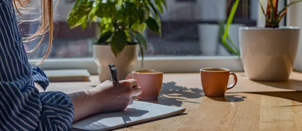 Mulheres mão closeup escrita com caneta no caderno ou planejador, tomando notas no café com xícara de café e plantas na mesa de madeira — Fotografia de Stock