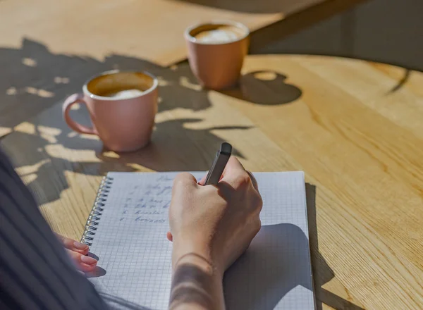 Mulheres mão escrever planos em notebook ou planejador sobre mesa de café de madeira com luz solar de janela e xícaras de café — Fotografia de Stock