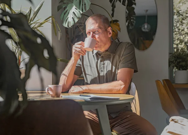 Reifer Mann sitzt an Holztisch in modernem Café mit Kaffeetasse zwischen Pflanzen — Stockfoto
