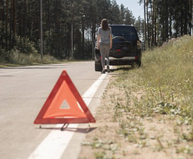 Arka plandaki arabaya giden ve acil durum onarım servisini bekleyen genç bir bayanla yol kenarında üçgen uyarı işareti.