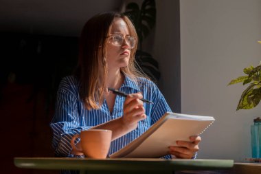 Kafede masada oturan düşünceli bir kadın, pencereden bakıyor, planlamacıyla planlar hakkında yazıyor ve düşünüyor.