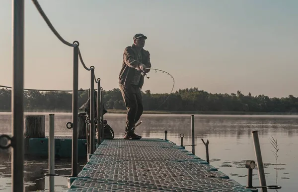 Pescatore pesca nella nebbia mattina. Uomo con asta di pesce appoggiato sul lago nella nebbia — Foto Stock