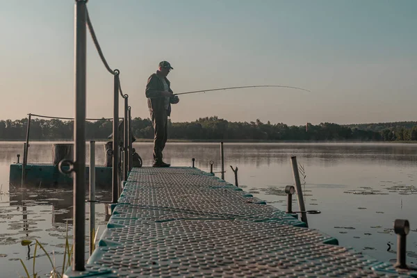 Pescatore pesca nella nebbia mattina. Uomo con asta di pesce appoggiato sul lago nella nebbia — Foto Stock