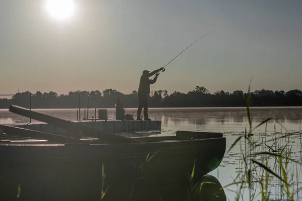 Pescatore pesca nella mattina presto serena. Uomo silhouette nera poggiante con asta sul fiume o lago con luce solare — Foto Stock