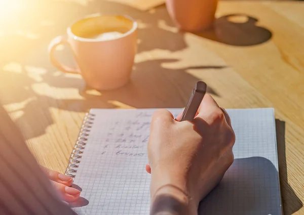 Planos de escrita à mão em notebook ou planejador sobre mesa de café de madeira com luz solar da janela e xícara de café, close-up — Fotografia de Stock