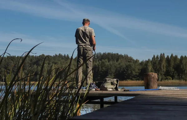 Pescatore su pontile di legno cattura pesci su asta bobina, in piedi in estate vicino al lago — Foto Stock
