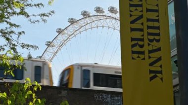 Londra, İngiltere - 1 Temmuz 2021: Demiryolu köprüsüne bakan tren arka planda Londra Gözü ve ağaç dalları ve ön planda bir Southbank Merkez tabelasıyla geçerken bir manzara