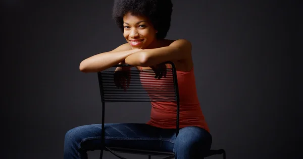 Leende svart kvinna sitter med armarna över baksidan av stolen — Stockfoto