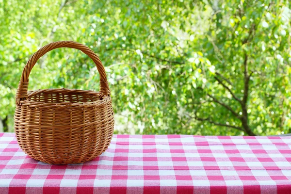 Empty basket on the table with checkered table cloth on a background of green trees. — Stock Photo, Image