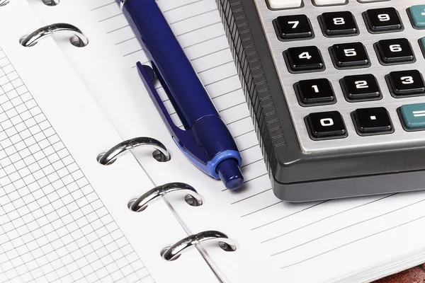 Açık defter üzerinde mavi kalem ve hesap makinesi yatıyor