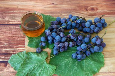 Kahverengi ahşap zemin üzerinde yapraklı mavi üzüm demetleri ve bir bardak şarap.