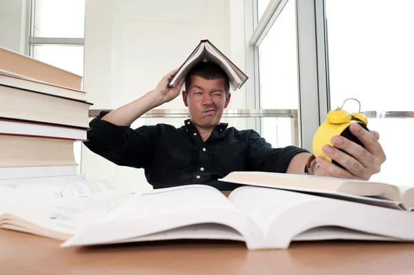 Closeup portrait of young man surrounded by tons of books, alarm clock, stressed from project deadline, study, exams. Negative emotion facial expression feelings, body language