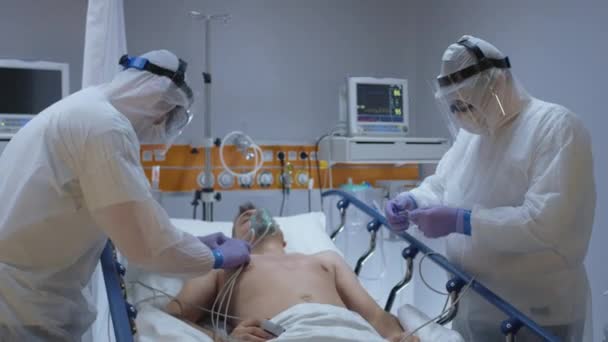 Lekarze w kombinezonach ochronnych Dołączenie EKG do pacjenta z koronawirusem — Wideo stockowe