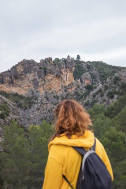 Woman from behind with a yellow jacket sits on a rock on top of a mountain in autumn with mist