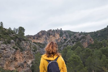 Woman from behind with a yellow jacket sits on a rock on top of a mountain in autumn with mist